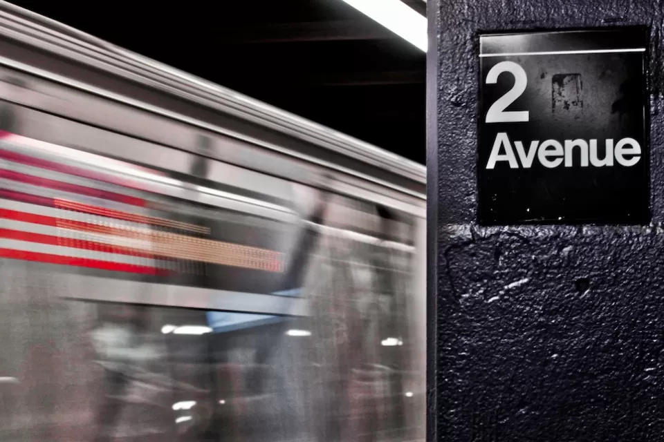 Close-up of 2 Avenue sign on column in NYC subway station with train passing by in background.